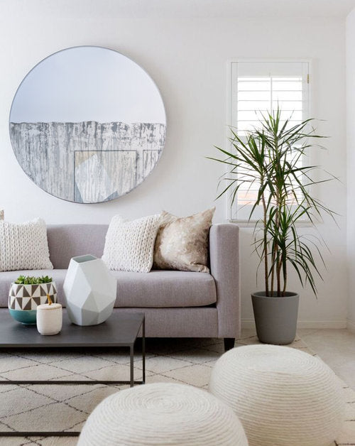 Circular mirror, mounted on wall, with grey mixed media painting on lower half. Displayed in living room above couch. Coffee table in front of couch and potted plant placed to the right of the couch with floor pillows in the foreground. 
