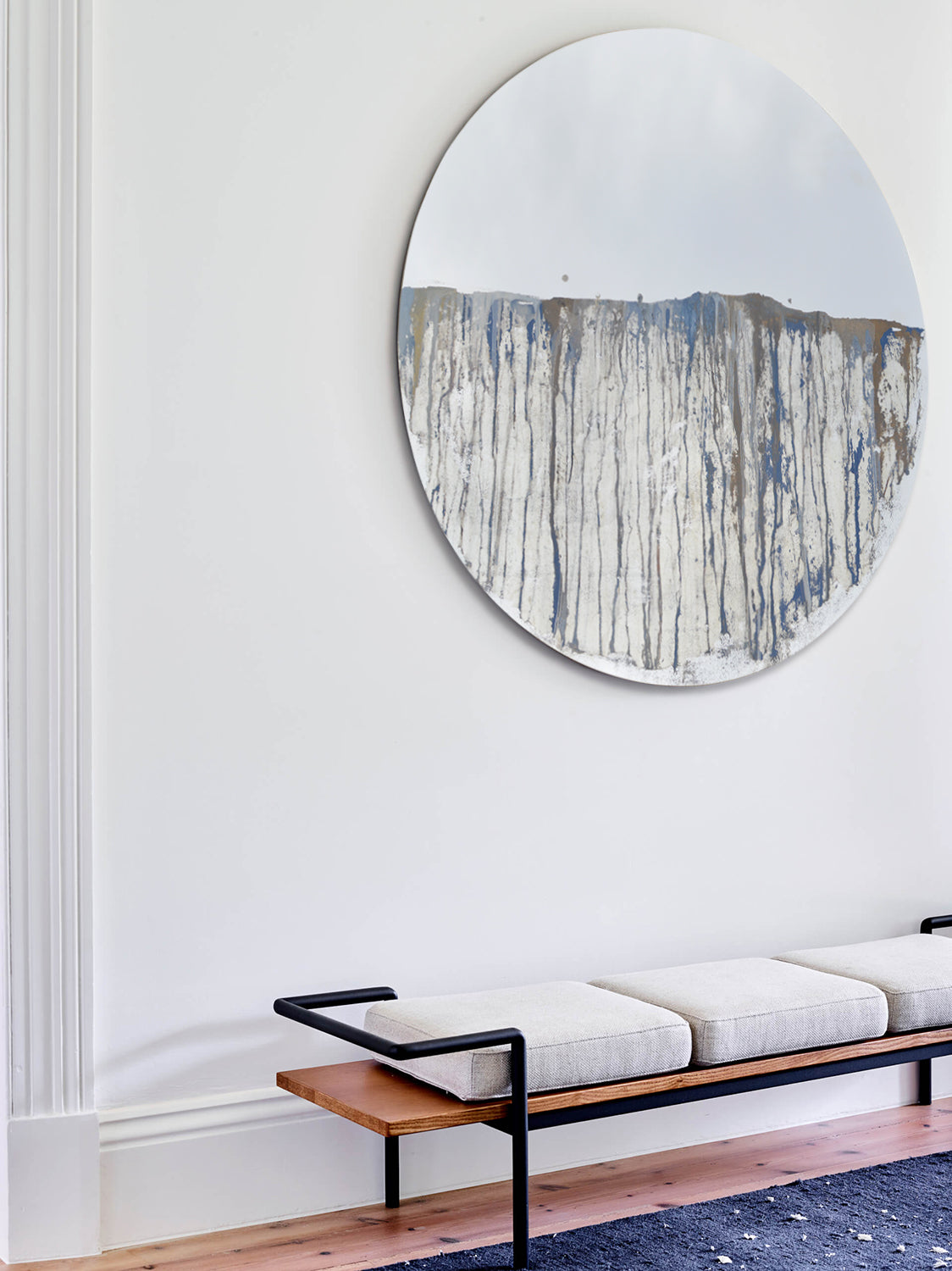 Circular mirror, mounted on wall, with blue and gold mixed media painting on lower half. Displayed above bench with seat cushions. 