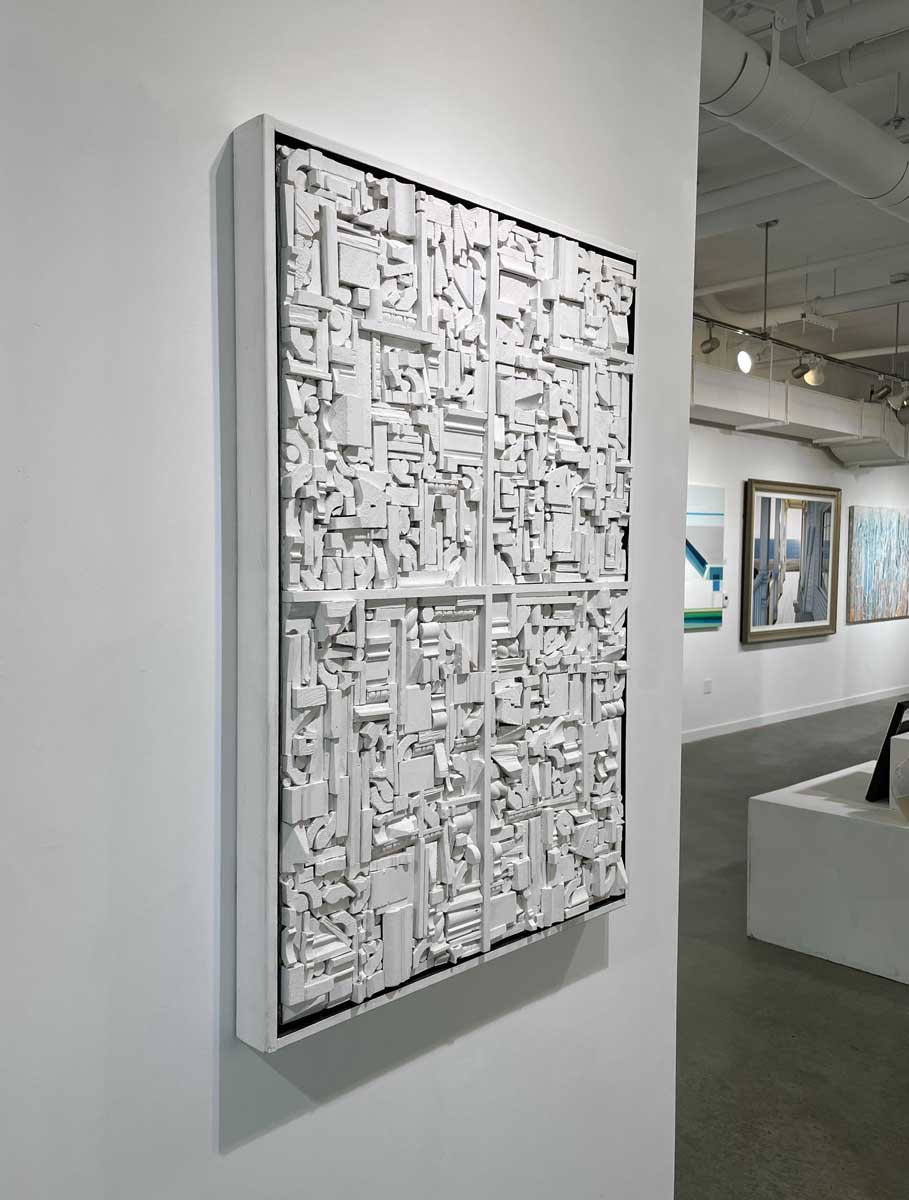A left side-angle view of a white geometric abstract wood construction in a white frame hangs on a white wall.