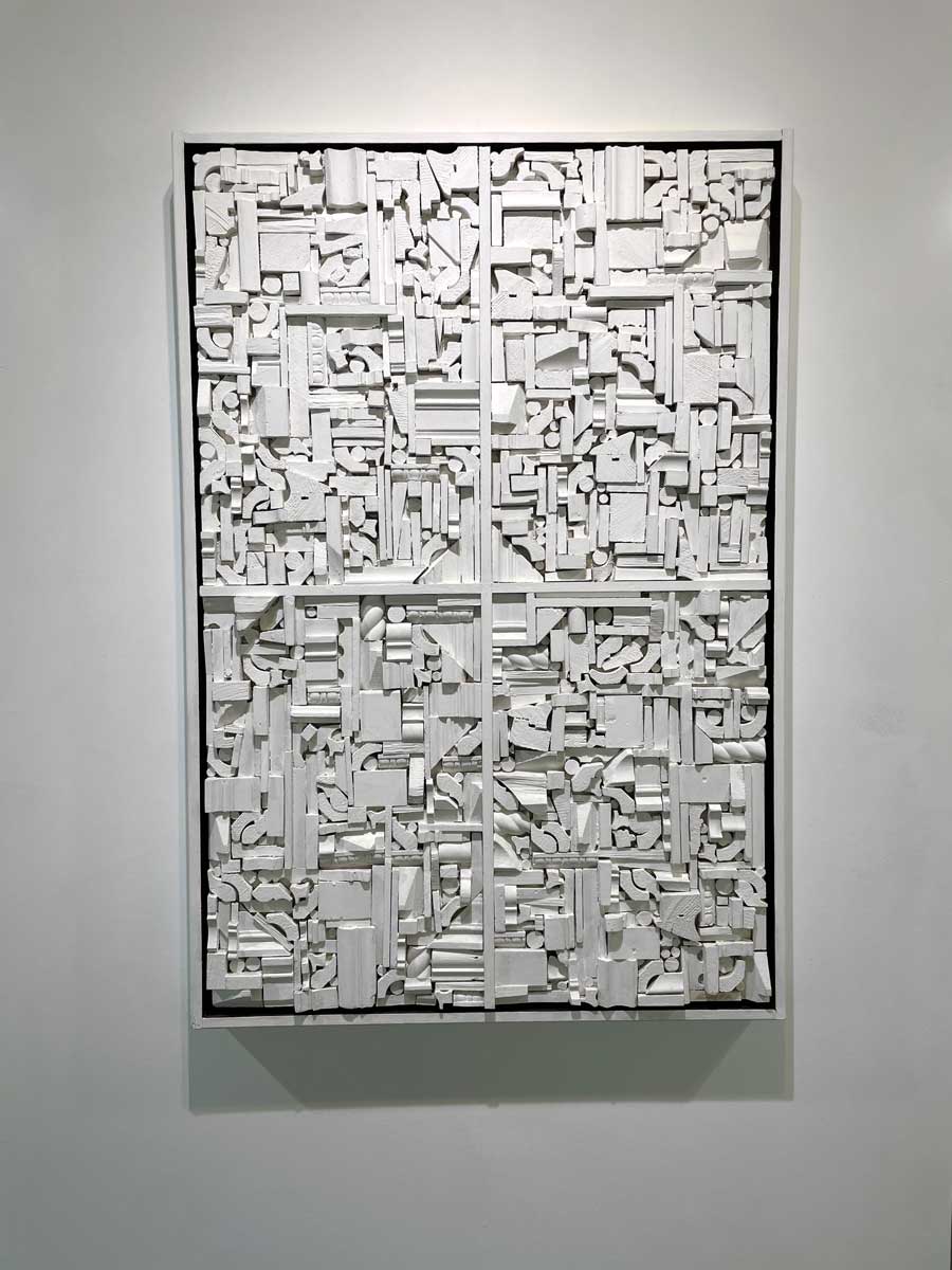A white geometric abstract wood construction in a white frame hangs on a white wall.