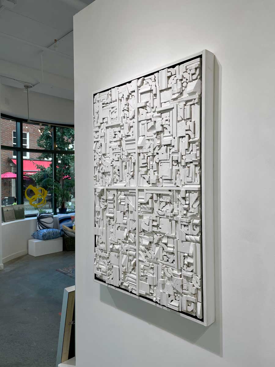 A right side-angle view of a white geometric abstract wood construction in a white frame hangs on a white wall.