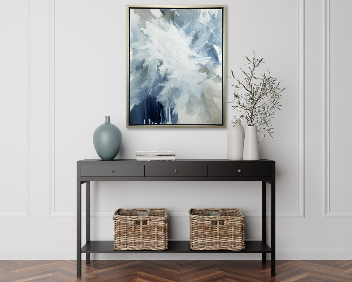 A white, blue and grey abstract print in a silver floater frame hangs on a white wall above a black console table with a muted teal vase, two white vases with greenery stems and two wicker baskets.