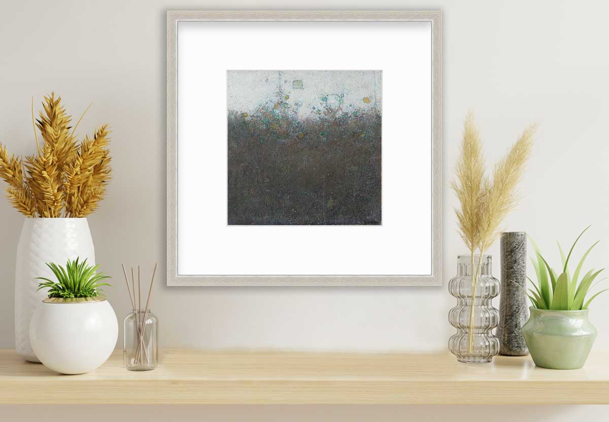 A dark olive green abstract landscape print in a silver frame with a mat hangs on a white wall above a wood surface with several vases with wheat and greenery.
