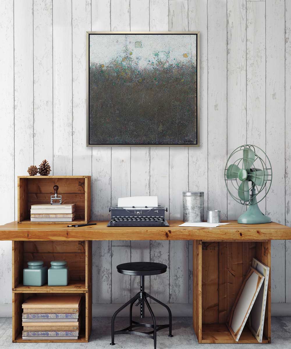 A dark green abstract landscape print in a silver floater frame hangs on a white wall above a natural wood desk with a typewriter, a green fan and accessories.