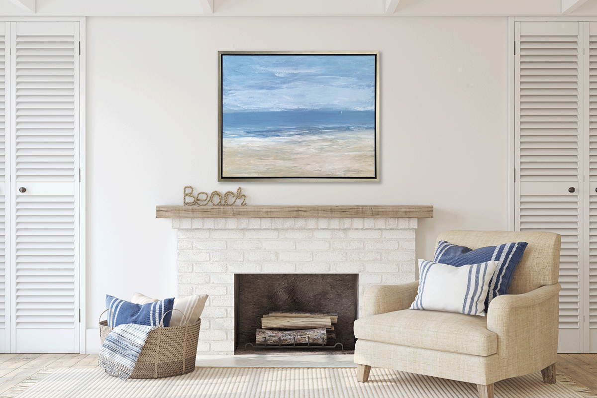 A blue, light blue, white and beige abstract seascape print in a silver floater frame hangs on a white wall above a white brick fireplace, a beige linen armchair with pillows and a woven basket with pillows.