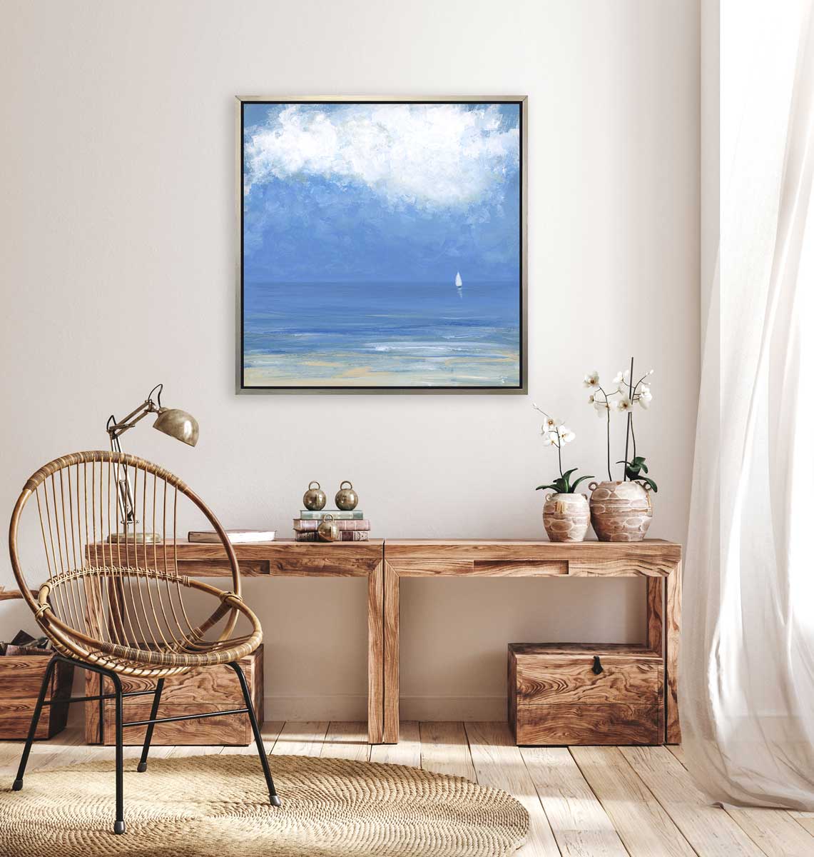 A blue, beige and white abstract seascape print with a small sailboat in a silver floater frame hangs on a white wall above a wood desk with books, a desk lamp and two potted white orchids.