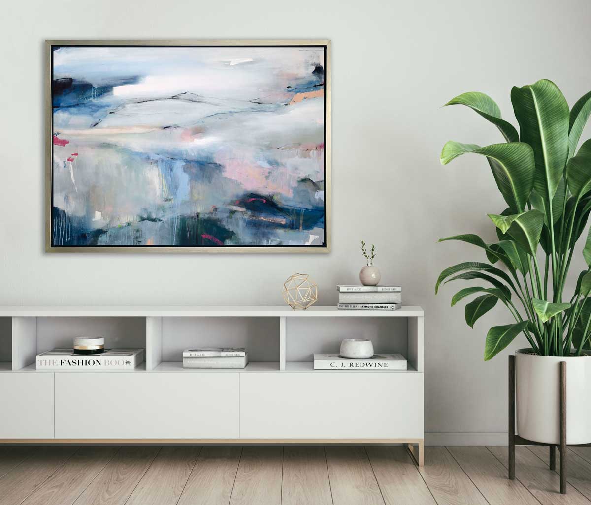 A blue, pink and white abstract print in a silver floater frame hangs on a white wall above a white sideboard with books, a gold wire sphere, a small round beige vase and a potted floor plant.