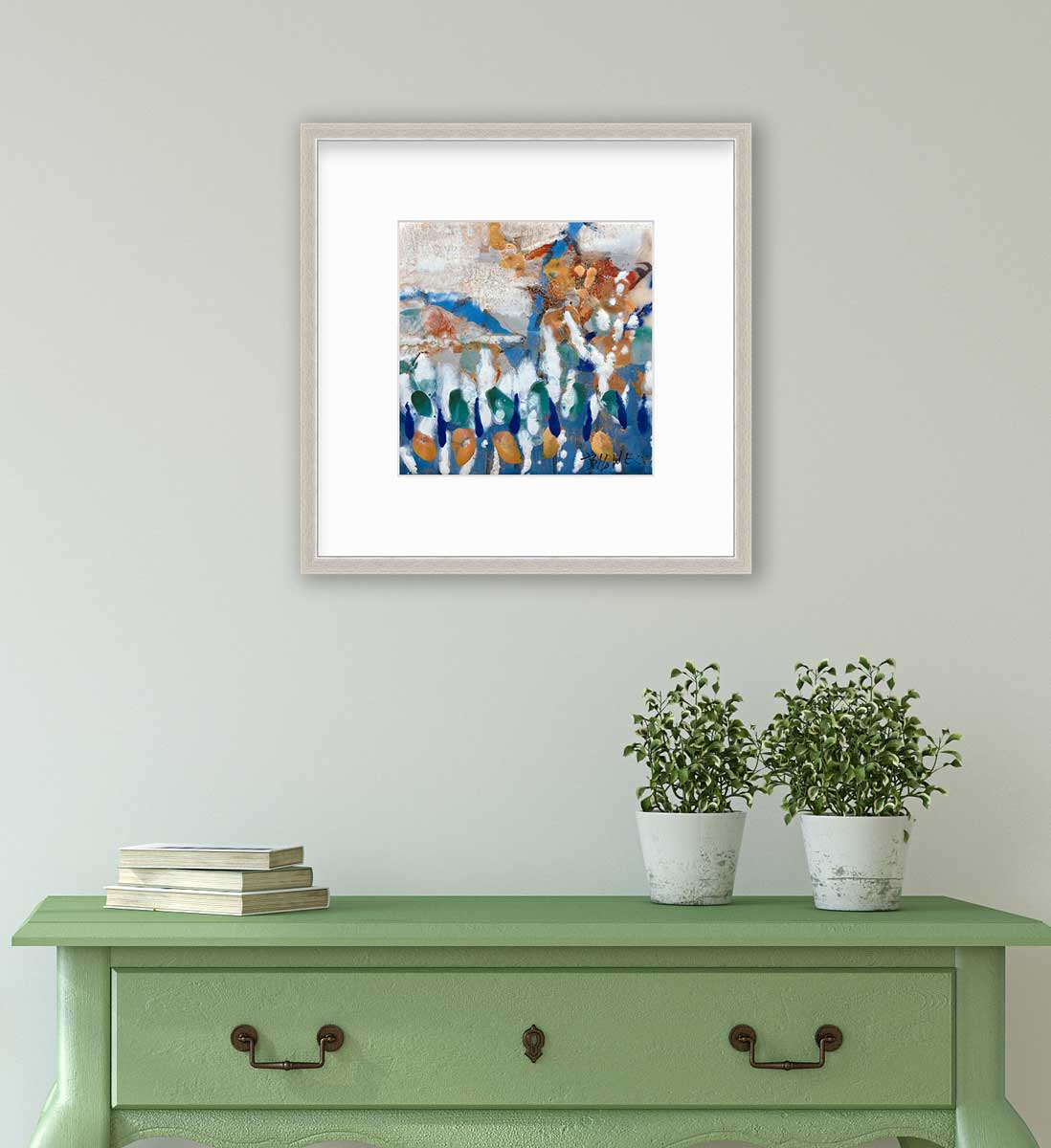 A multicolored abstract print in a silver frame with a mat hangs on a white wall above a green console table with a stack of books and two potted plants.