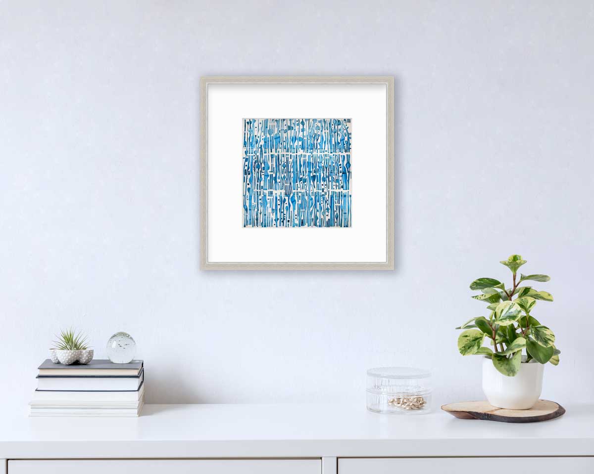 A blue and white abstract print in a silver frame with a mat hangs on a white wall above a white surface with a stack of books, a round clear container with gold accessories and a potted plant.