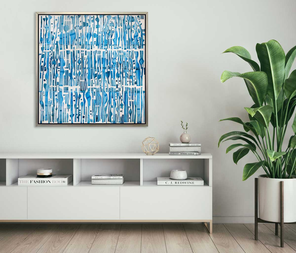 A blue and white abstract print in a silver floater frame hangs on a white wall above a white cabinet with books, a gold wire sphere, a small vase with two green stems and a potted floor plant.