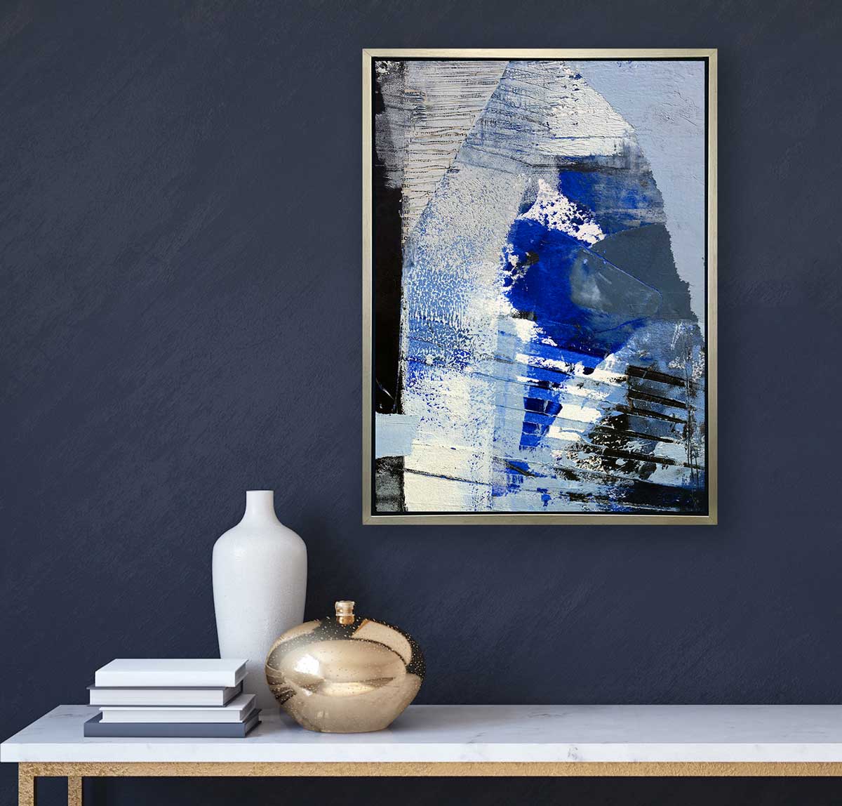 A blue, white and black abstract print in a silver floater frame hangs on a blue wall above a white marble console table with a stack of books, a white vase and a gold vase.