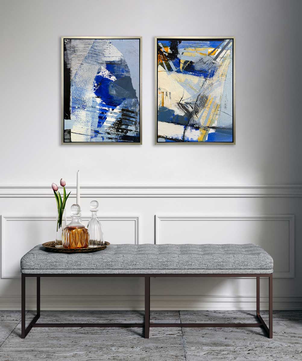 Two blue, black, white and grey abstract prints in silver floater frames hang side by side on a white wall above a grey tufted bench with a tray of glass decanters, a glass vase with a pink tulip and a white tapered candle.