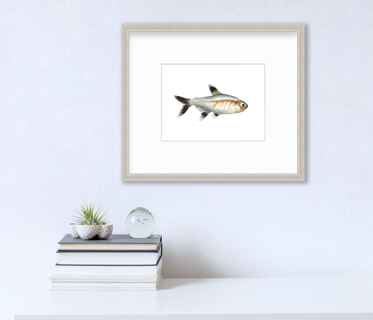 A black, silver and brown fish print in a silver frame with a mat hangs on a white wall above a white surface with a stack of books, a clear round paperweight and a small succulent decoration.