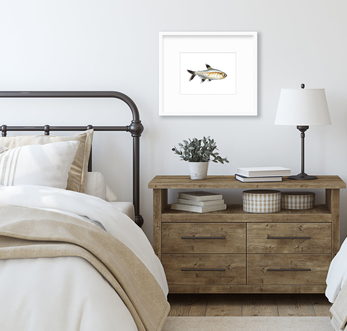 A black, silver and brown fish print in a silver frame with a mat hangs on a white wall above a wood nightstand with stacks of books, a table lamp, a potted plant and a black wrought iron bed with neutral bedding.