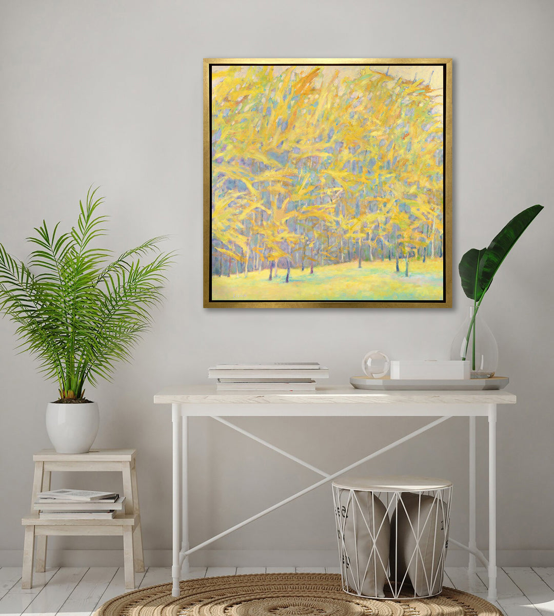 A yellow and blue abstract tree landscape print in a gold floater frame hangs on a white wall above a white desk with a stack of books, a tray that has a glass vase with green leaf stems and a white step stool with a potted plant.