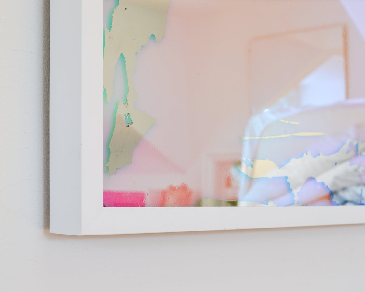 A close up view of a corner of the pink, yellow and orange dichroic film art mirror in a white frame hangs on a white wall.