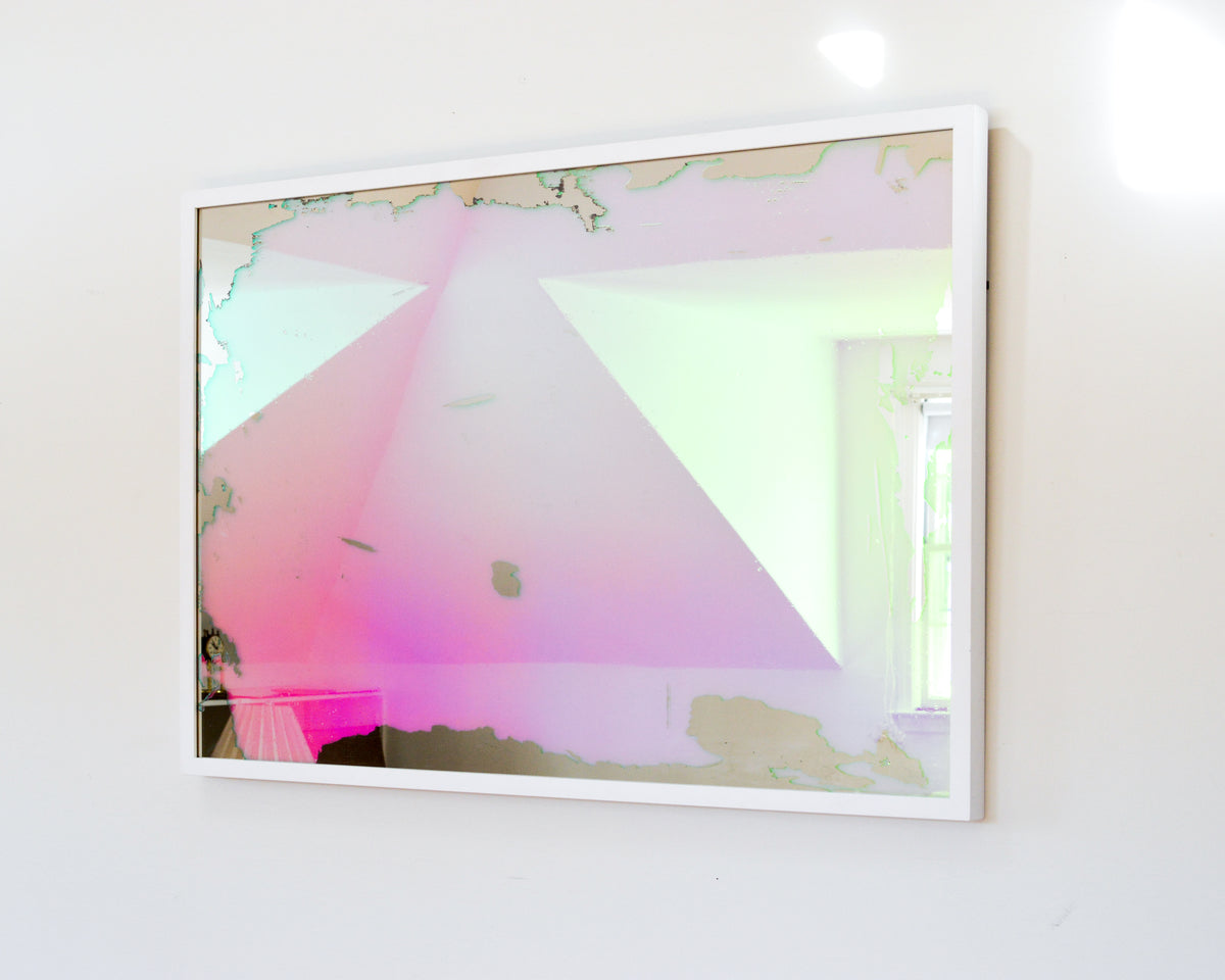 A right side-angle view of a pink, yellow and orange dichroic film art mirror in a white frame hangs on a white wall.