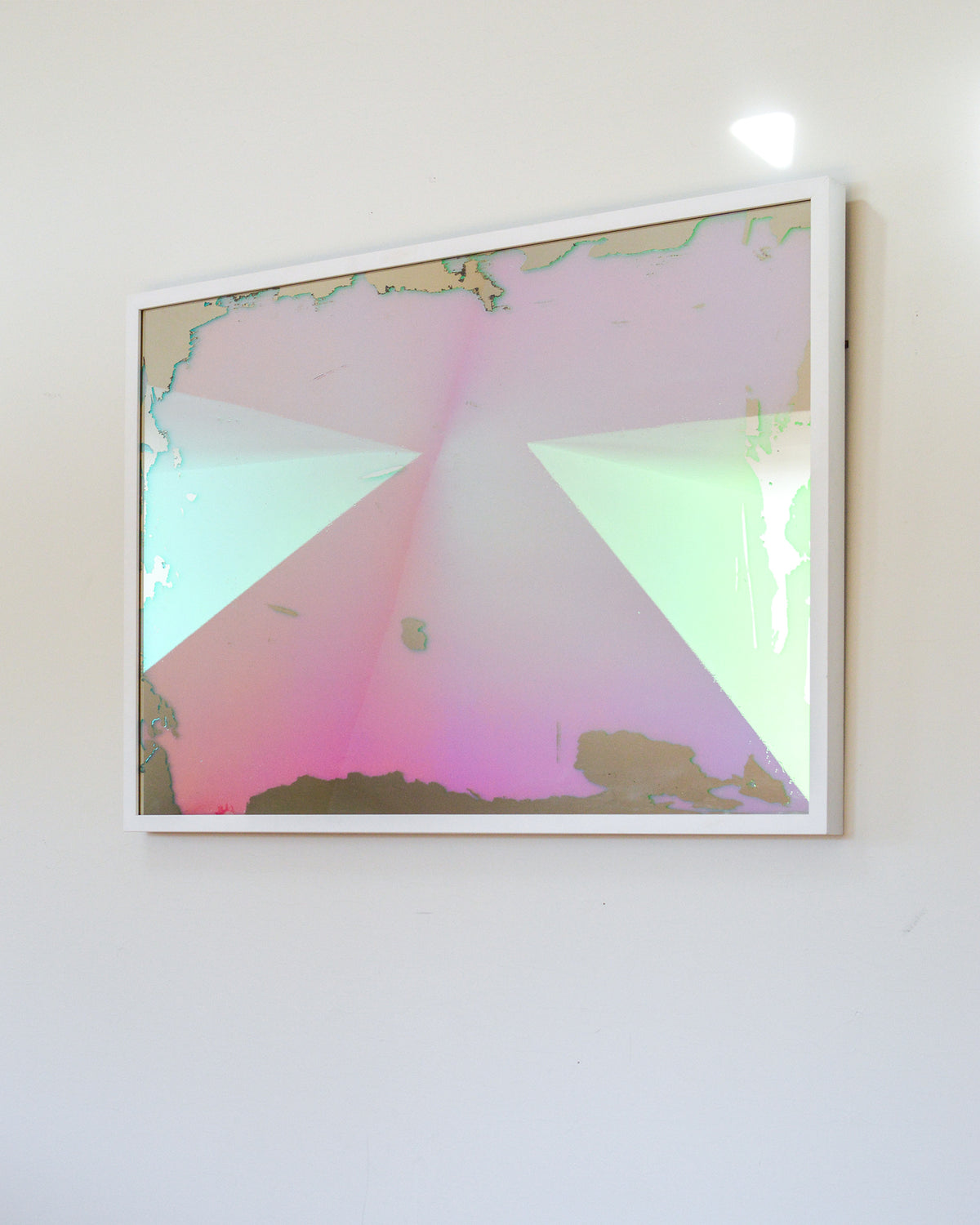 A right side-angle view of a pink, yellow and orange dichroic film art mirror in a white frame hangs on a white wall.