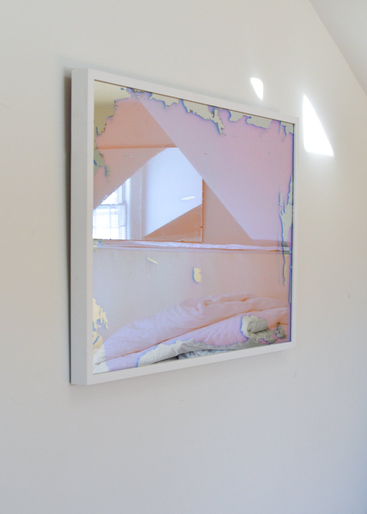 A left side-angle view of a pink, yellow and orange dichroic film art mirror in a white frame hangs on a white wall.
