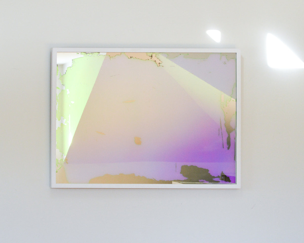 A pink, yellow and orange dichroic film art mirror in a white frame hangs on a white wall.