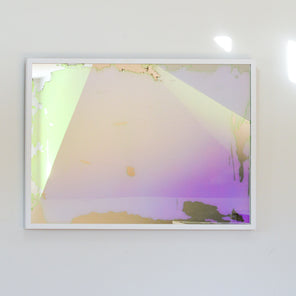 A pink, yellow and orange dichroic film art mirror in a white frame hangs on a white wall.