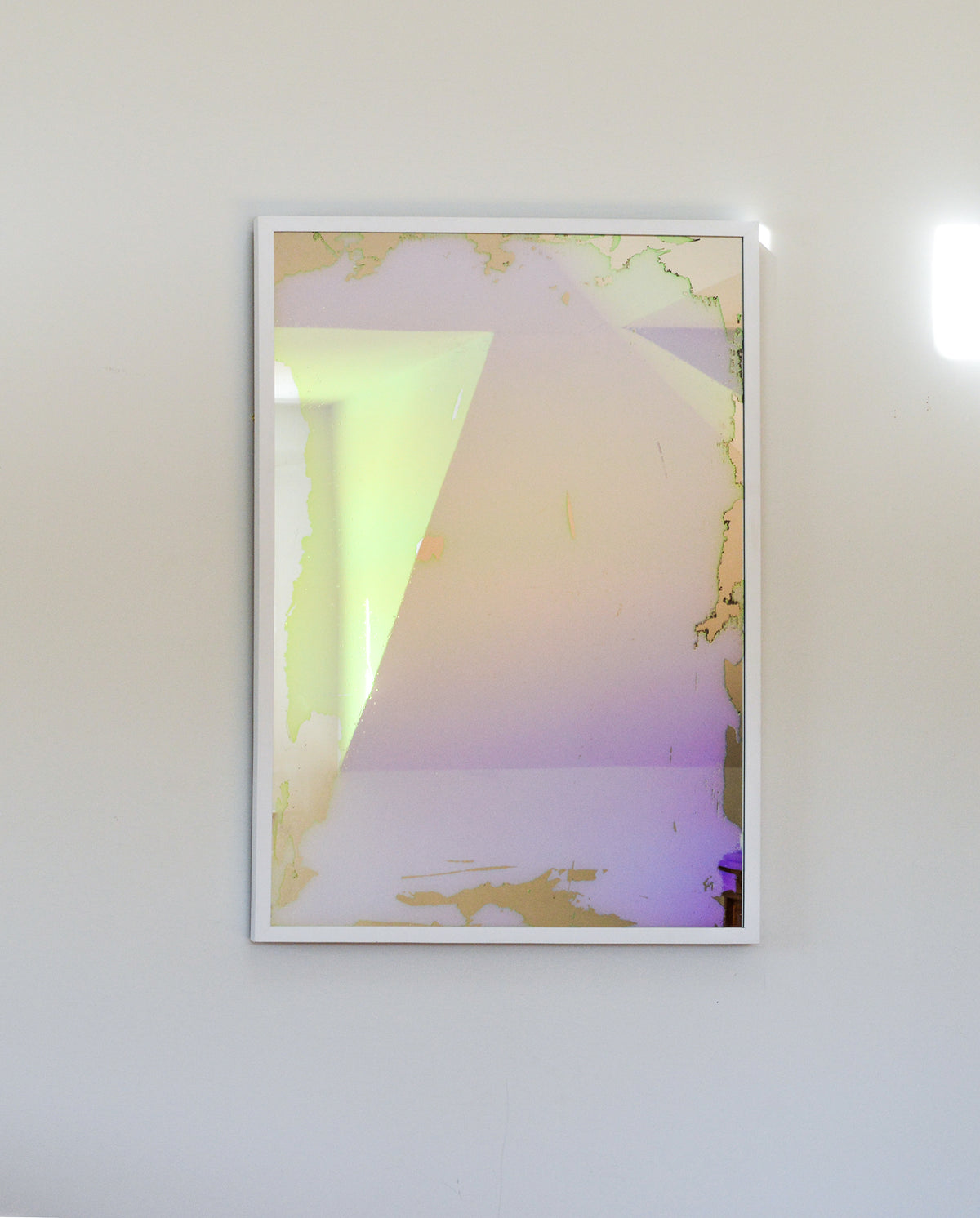 A pink, yellow and orange dichroic film art mirror in a white frame hangs vertically on a white wall.