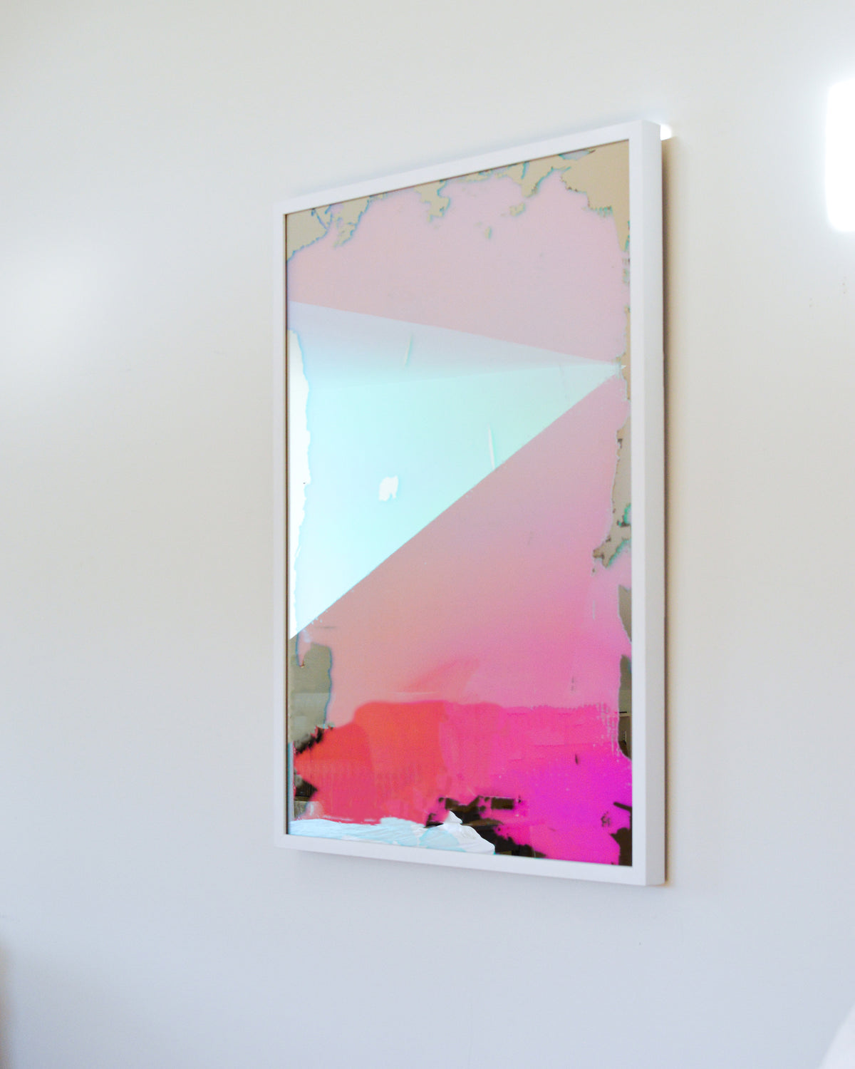 A right side-angle view of a pink, yellow and orange dichroic film art mirror in a white frame hangs vertically on a white wall.