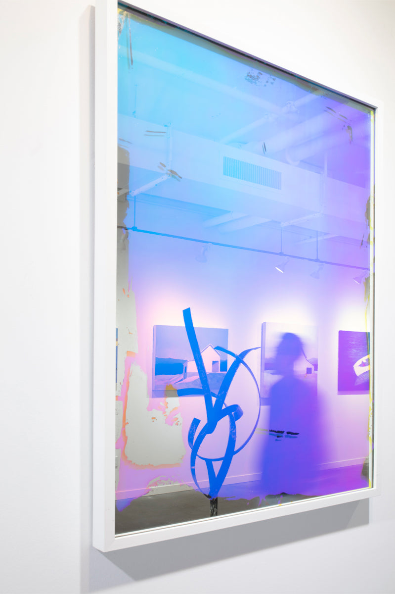 A multi-colored fine art mirror hangs on a white wall from an angled view, with reflections of the room and a blurred image of a person passing by. 