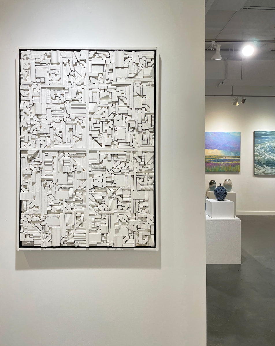 A white geometric abstract wood construction in a white frame hangs on a white wall in an art gallery with paintings on the wall in the background. 