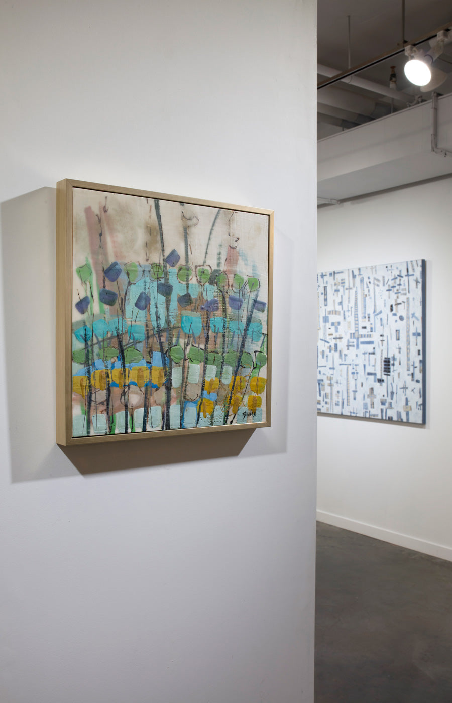 An abstract floral painting framed in a gold frame hangs on a white wall with a blue and white abstract painting hanging on a wall adjacent to it. 