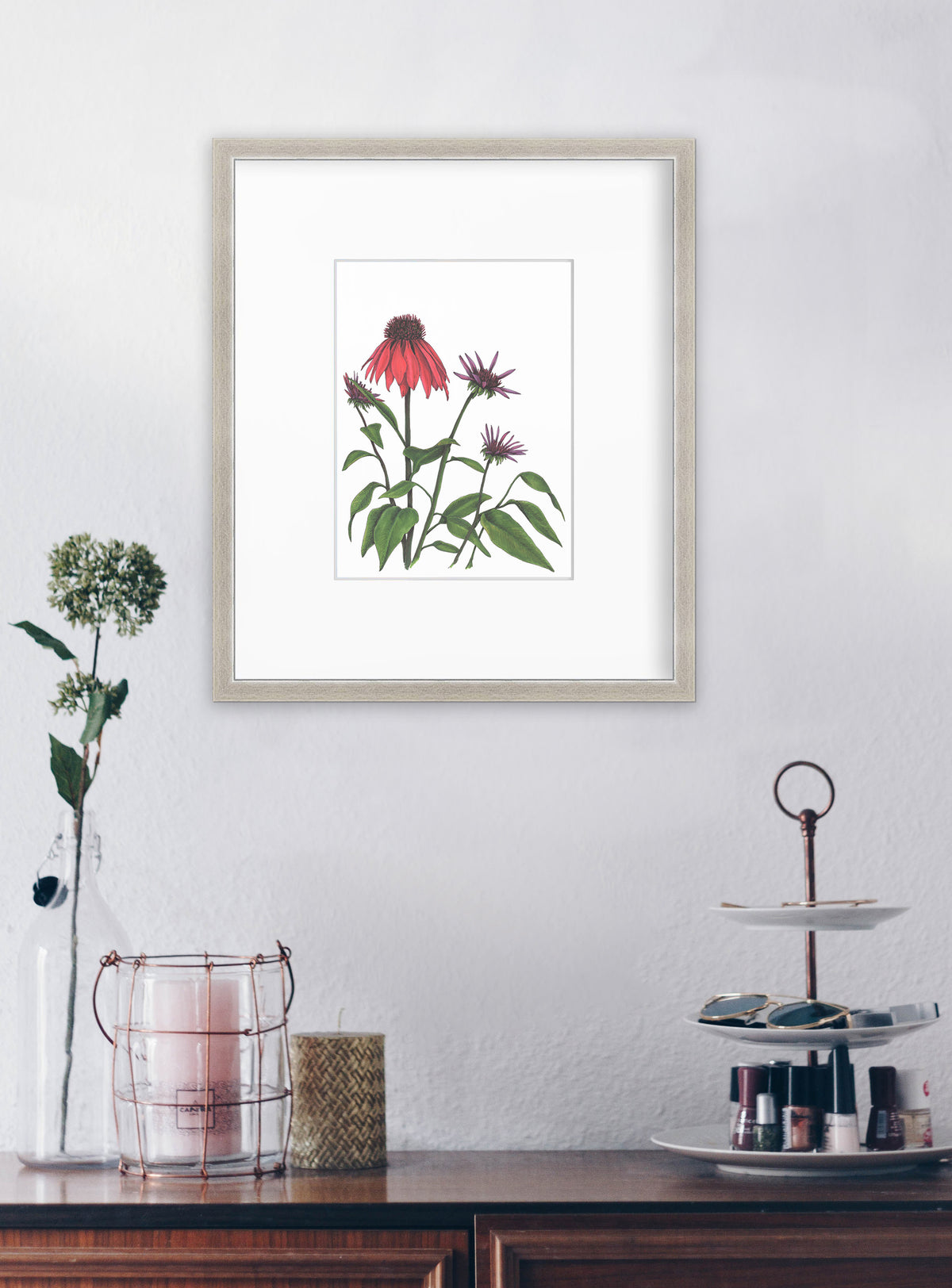 Red and purple flowers with green stems and leaves print in a silver frame with a mat hangs on a white wall above a wooden console with a candles and tray for holding nail polish. 