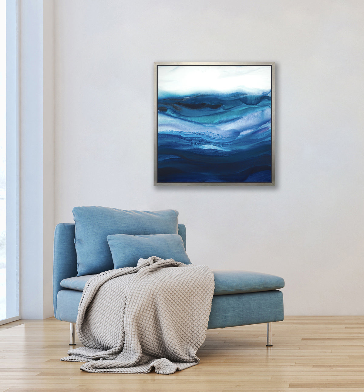 A blue, teal and white abstract print in a silver floater frame hangs on a white wall above a light blue armless chair with a cream colored throw blanket.