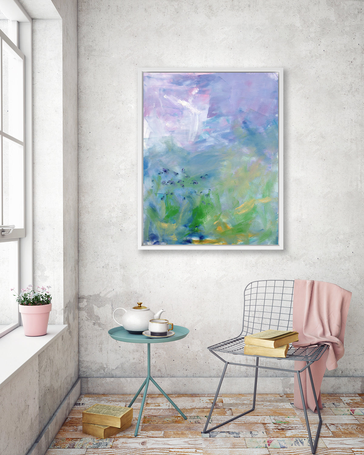 A blue, purple and green abstract print in a white floater frame hangs on a concrete wall above a grey wire chair with a book and a pink throw blanket, a stack of books on the floor and a small teal side table with a white tea set.