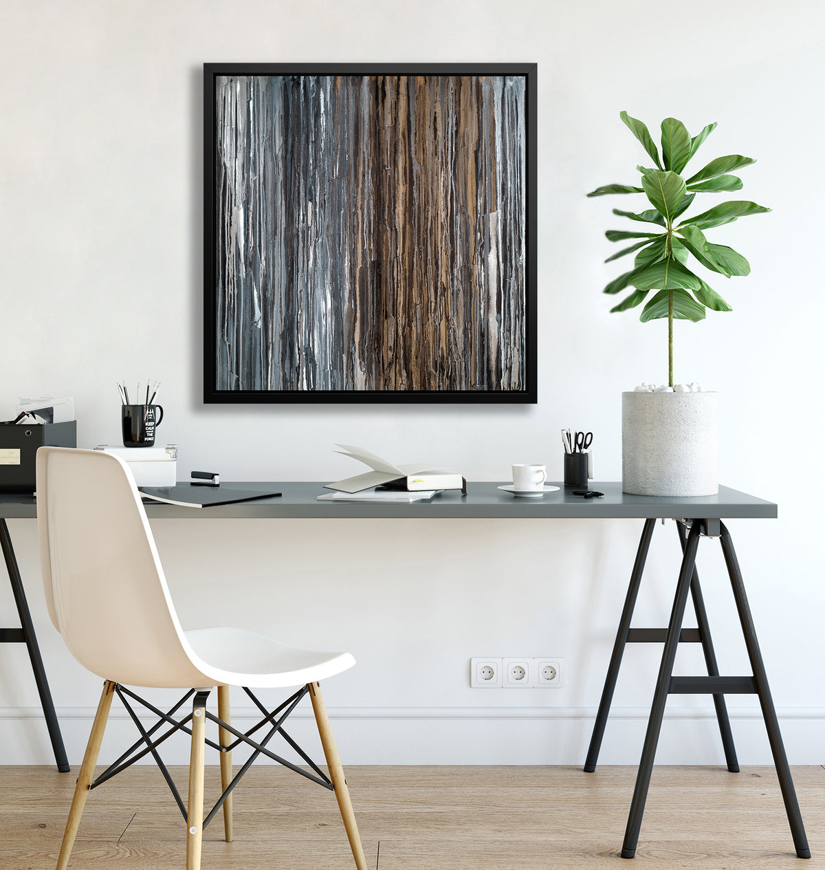 A brown, copper, white, grey and black abstract print in a black floater frame hangs on a white wall above a grey desk with open books, black coffee mugs with pencils and a potted plant.
