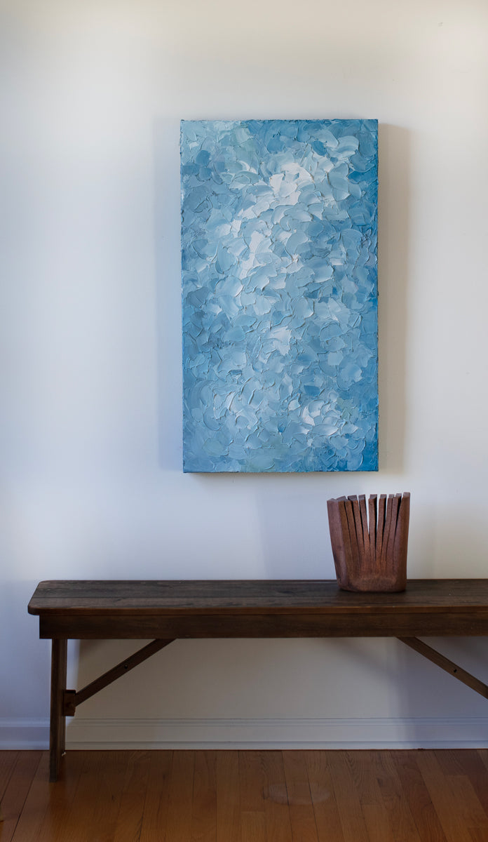 A blue and white abstract painting by Teodora Guererra hangs vertically on a white wall above a dark wood bench with a ceramic sculpture accent.