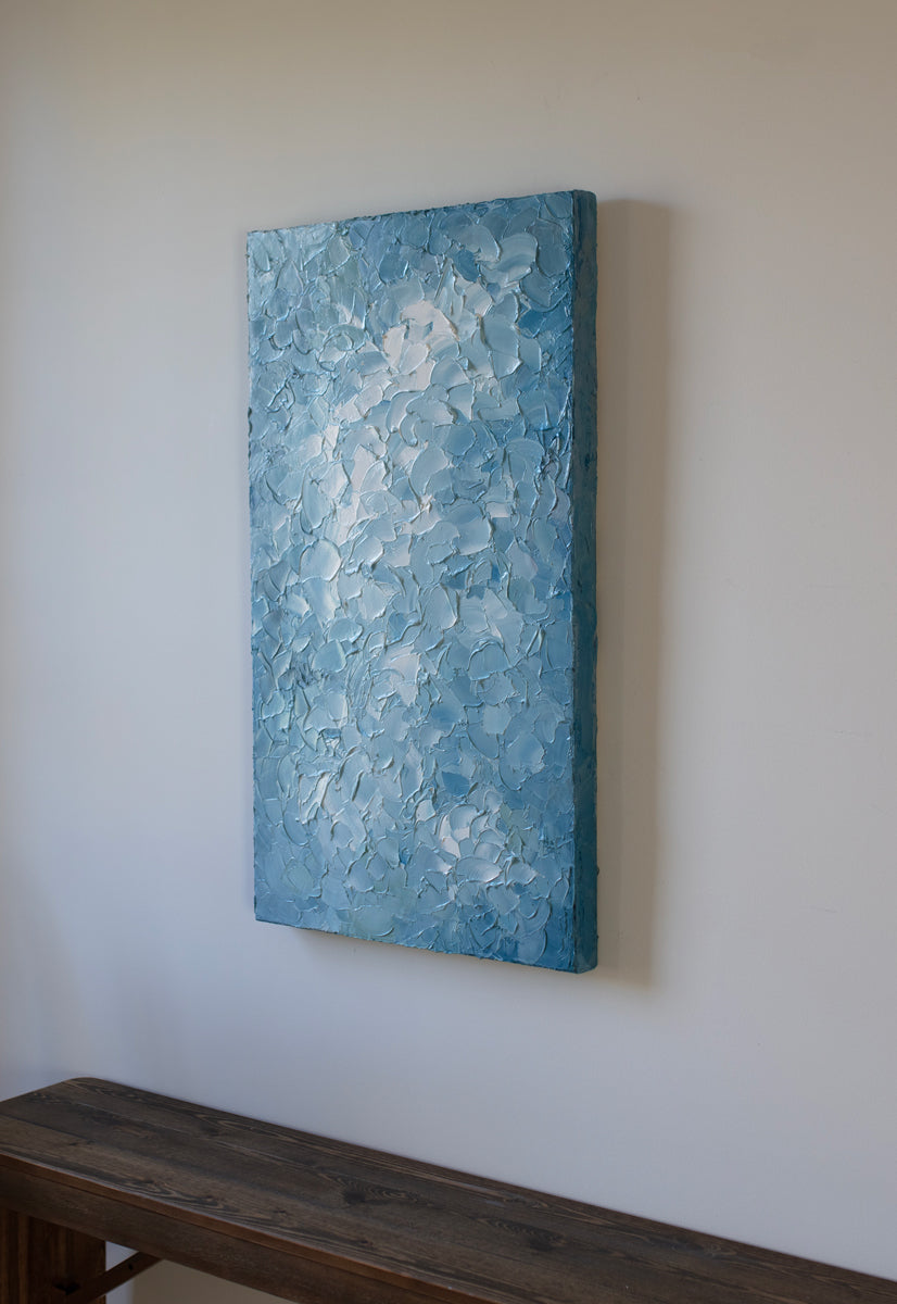 A right side-view of a blue and white abstract painting by Teodora Guererra hangs on a white wall above a dark wood bench.