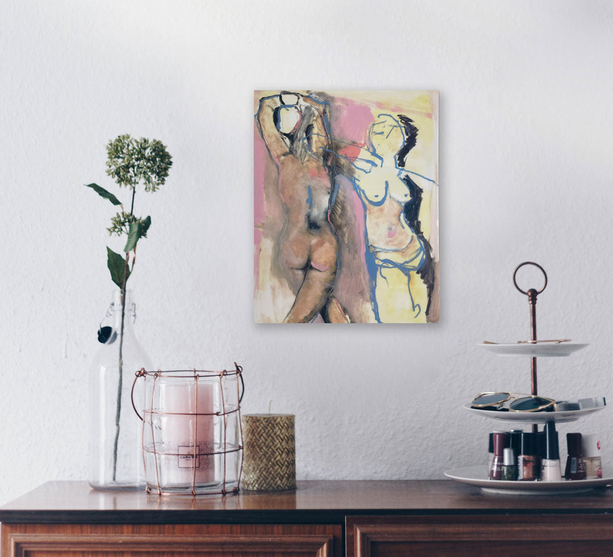 A pink and yellow abstract figurative print hangs above a wooden dresser with candles and a tiered accessories tray. 