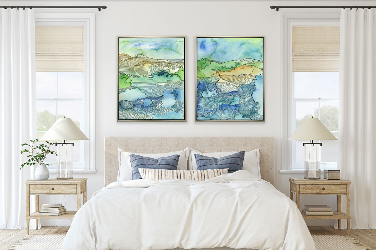 Two landscape illustration prints hang on a white wall above a neutral and blue bed, wooden end tables, and modern lamps. 