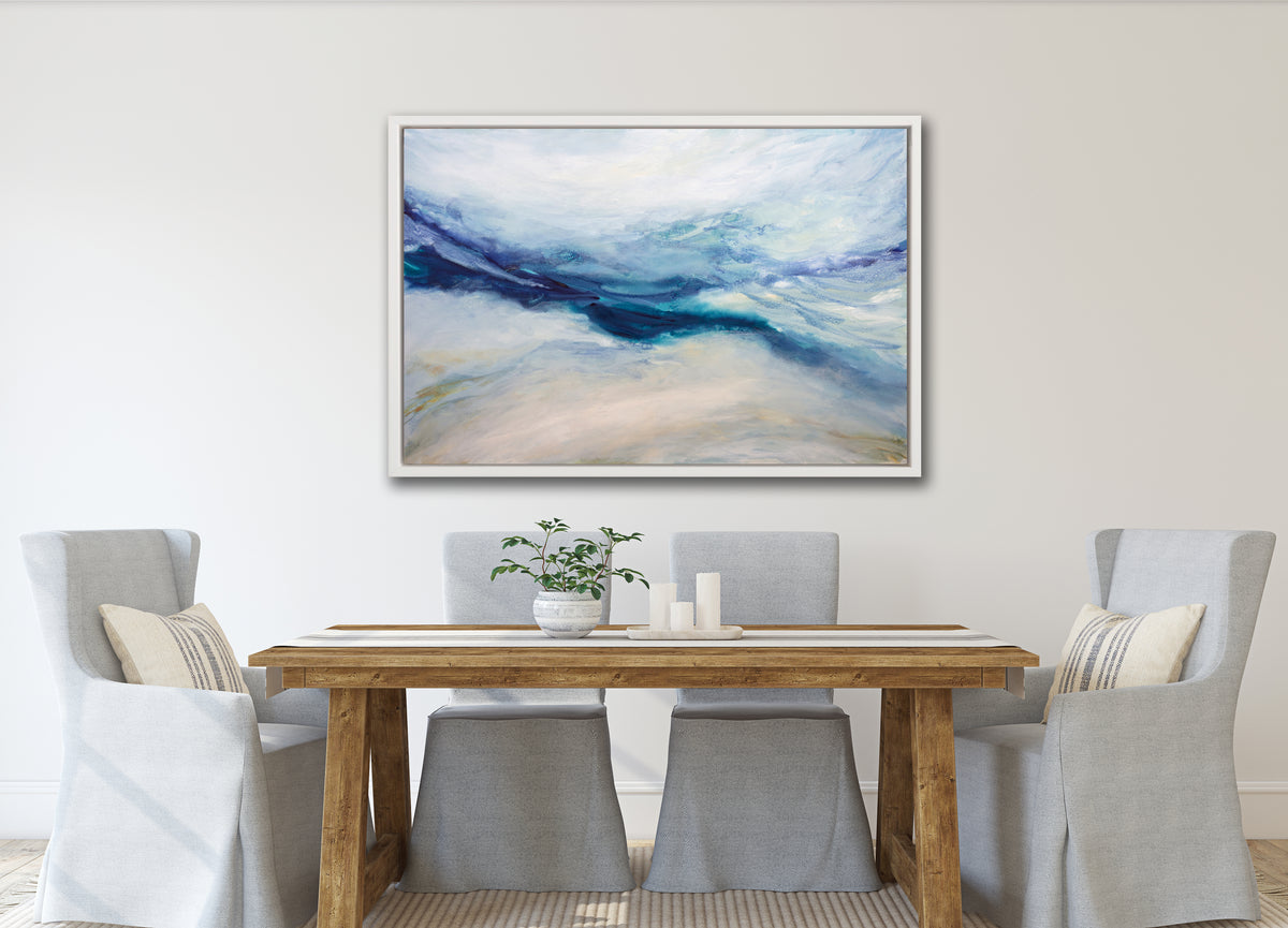 A white, blue, purple and beige abstract print in a white floater frame hangs on a white wall above a wood table with four grey slipcovered chairs.