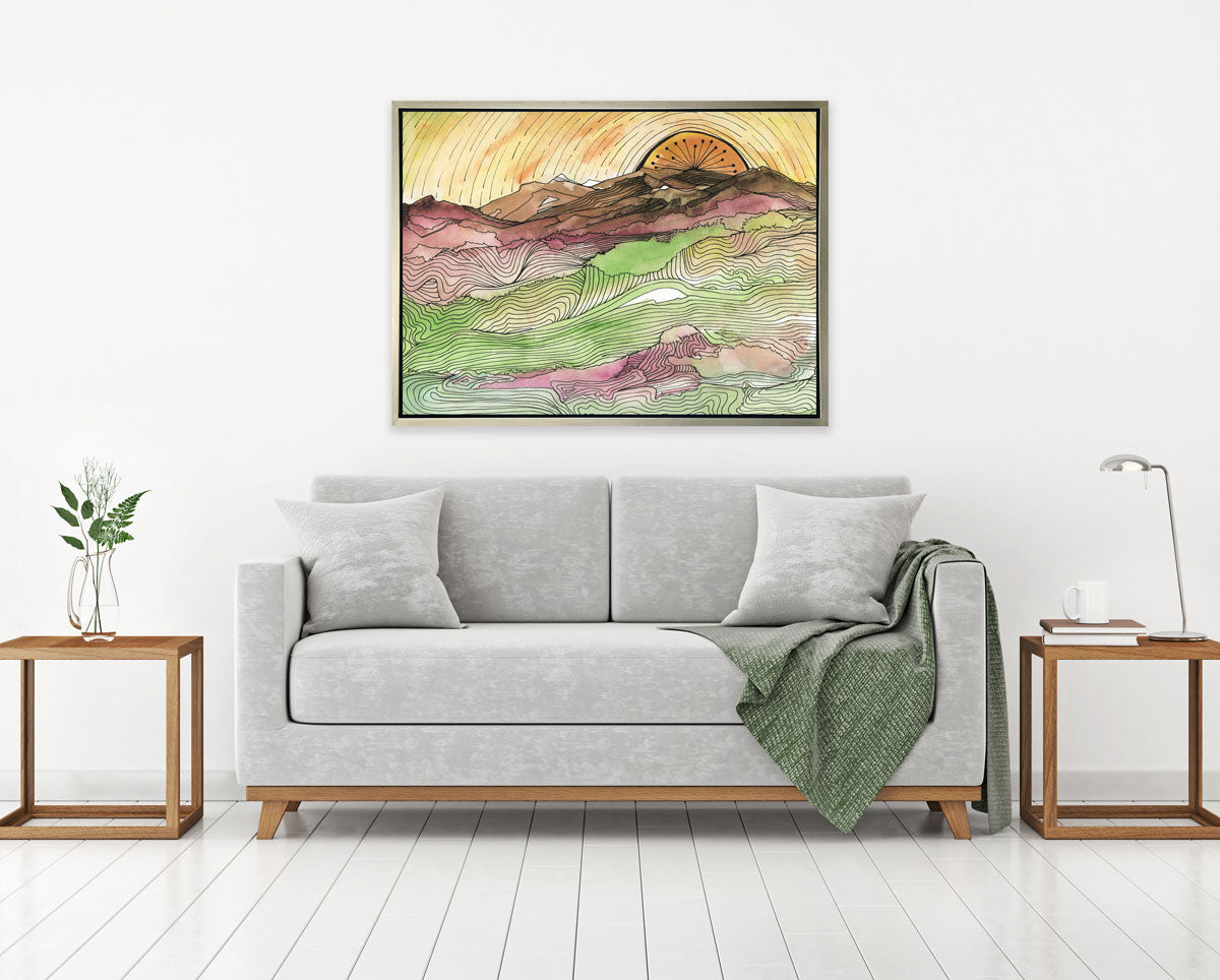 A yellow, brown, green and dark rose abstract landscape print with black outlines in a silver floater frame hangs on a white wall above a grey sofa with pillows and a green throw blanket and two wood side tables.