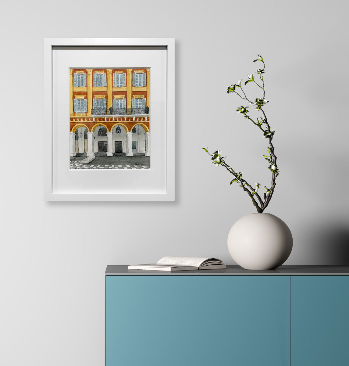 An orange and yellow architectural painting in a white frame hangs on a light grey wall above a turquoise dresser and modern white table vase with flowers, and open book, 