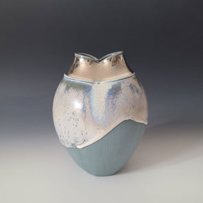 A blue textured sculptural vessel with light, multicolored glaze and a white mouth sitting on a gradient surface. 
