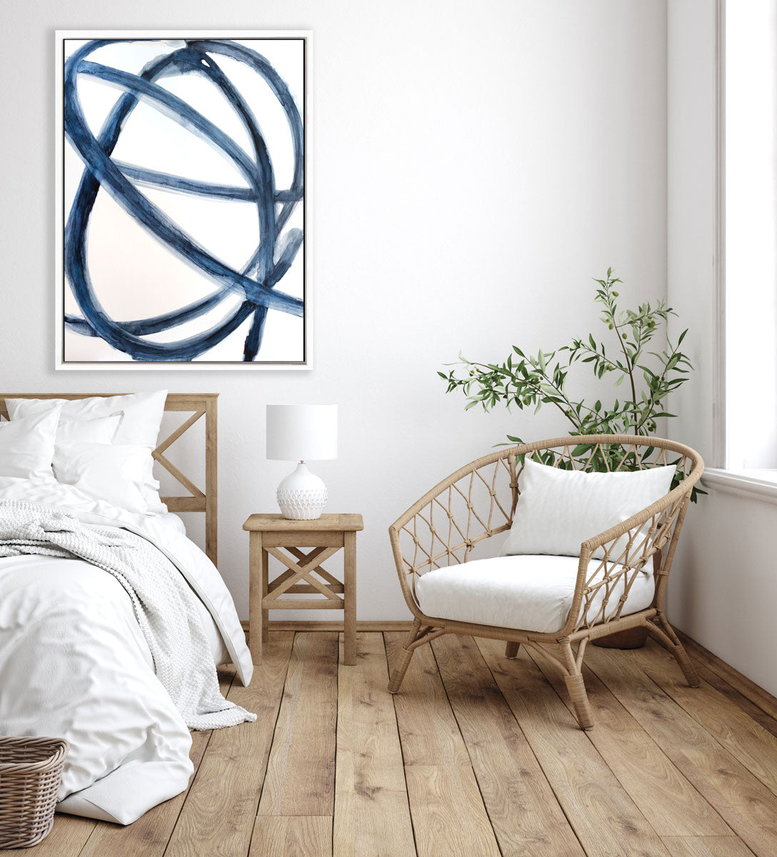 A blue and white abstract print hangs on a white wall above a wooden bed, end table, wicker chair, and floor plant. 