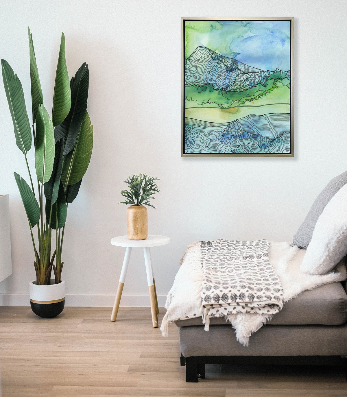 A blue and green landscape illustration print hangs on a white wall above a soft grey chair, a white end table with a potted plant, and a large floor plant. 
