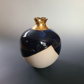 A white and dark blue ceramic sculptural vessel with a gold mouth sits on a grey gradient surface. 