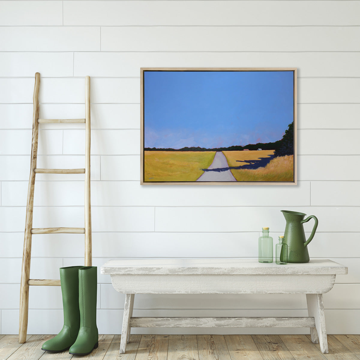 A blue and yellow-green abstract landscape print, framed in a white oak frame hangs on a white paneled wall above a white bench, green rain boots, watering can, and a ladder. 