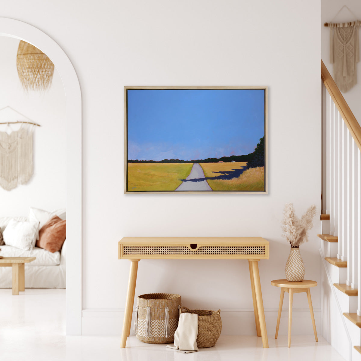 A blue and yellow-green abstract landscape print, framed in a white oak frame hangs on a white wall above a light wood console table, and between the entry to a living room and a stairwell. 