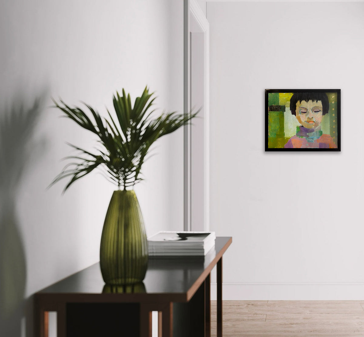 A green abstract portrait painting of a child with dark hair hangs on a white wall, near a wooden console table with a green vase and plant and table top book decor. 