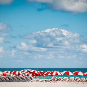 A photograph by Peter Mendelson of red and white striped umbrellas and lounge chairs on a beach beneath a cloudy blue sky. 