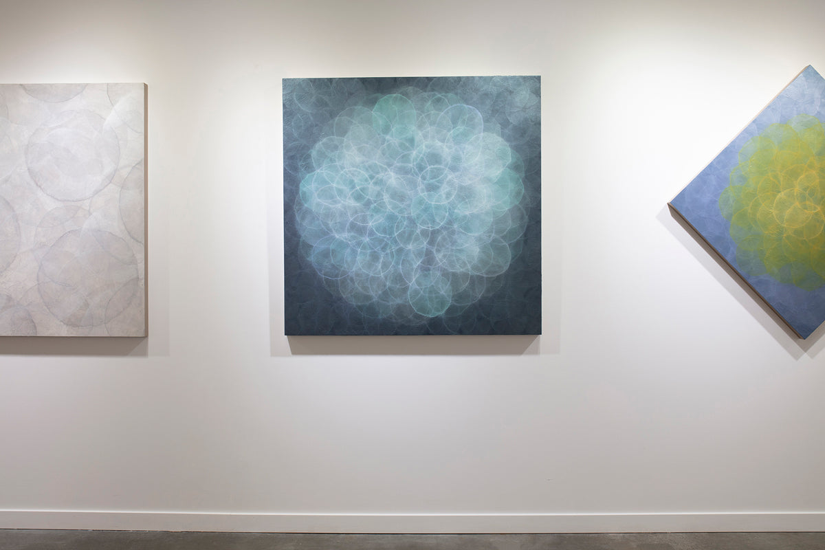 A blue, teal, light lavender and white abstract geometric painting hung on a white wall in dark lighting.